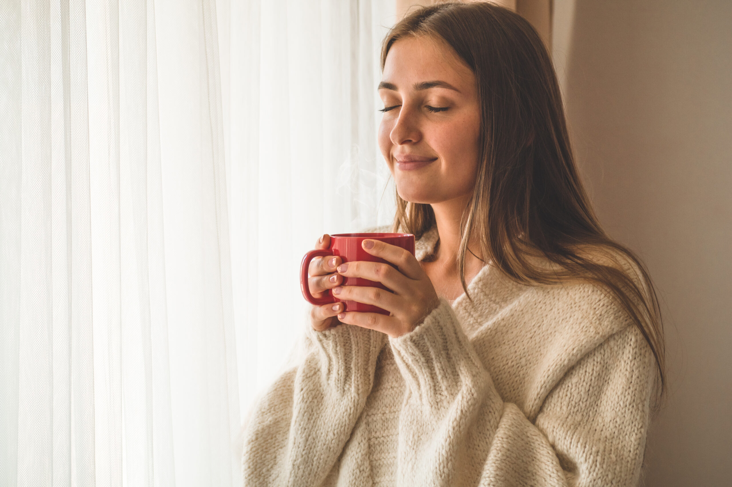 Frau hält entspannt eine warme Tasse am Fenster und genießt eine kurze Pause für mehr Wohlbefinden im Alltag.